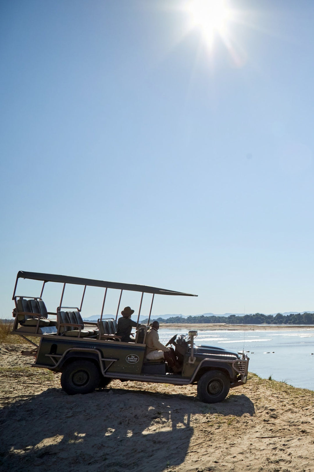 Jeep safari vehicle on a riverbank with wildlife in the distance under a clear blue sky.